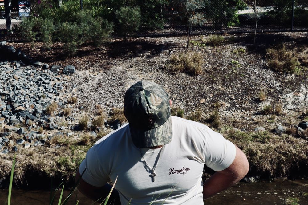 Bush Camo hat worn at a creek bank with the Ecru tee, seen from behind