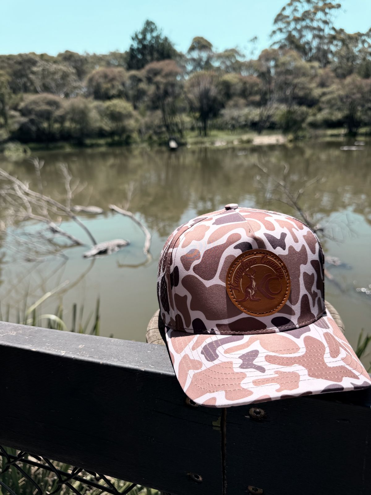 Bush Camo hat with leather Kangalope patch resting on a fence rail above a billabong