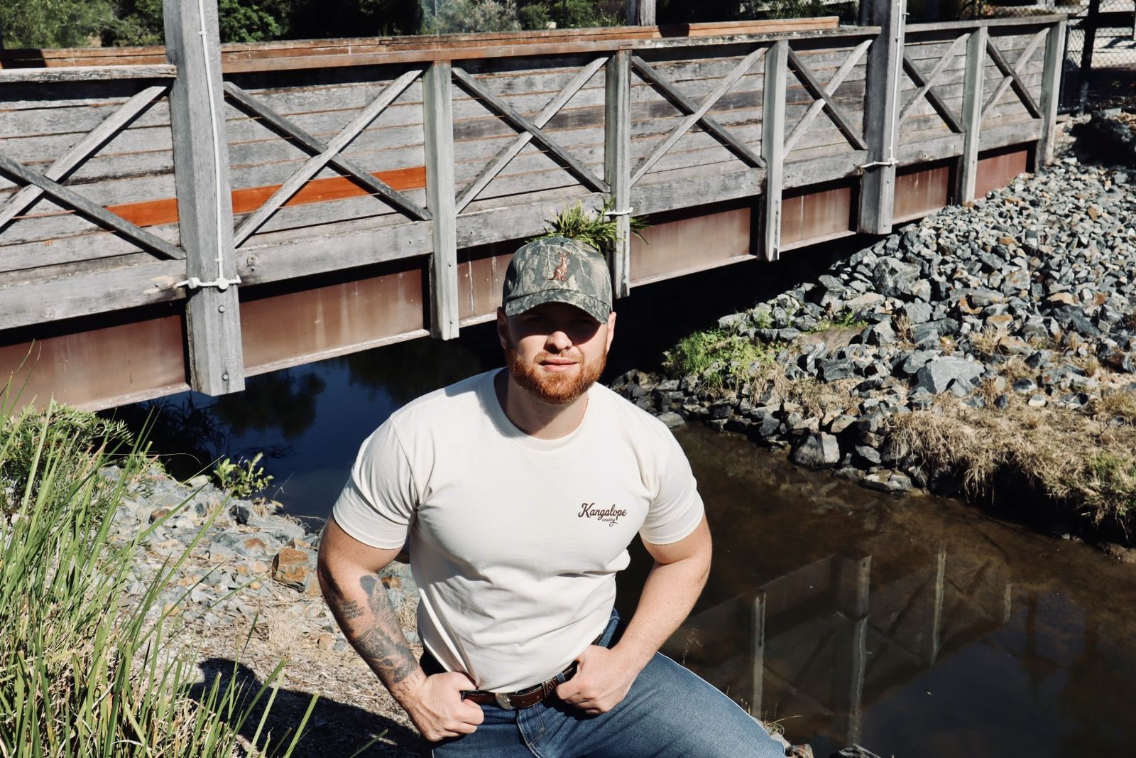 Front of the Ecru tee worn with a camo hat, standing on a timber bridge over a creek