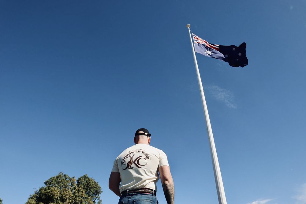 Back print of the Ecru tee beneath a flying Australian flag against blue sky